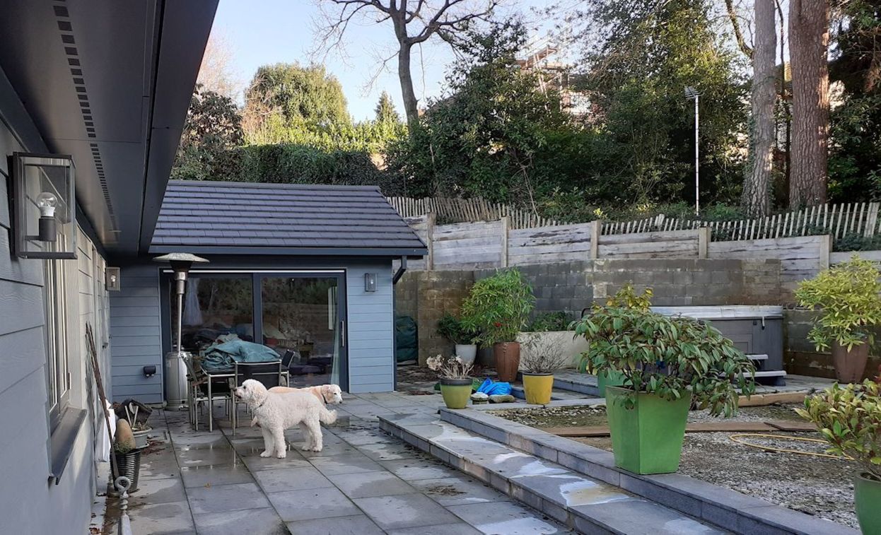 A sloping garden design in Hindhead, Surrey, with retaining walls, an outdoor kitchen, a waterfeature, built-in seating, and terraced levels.