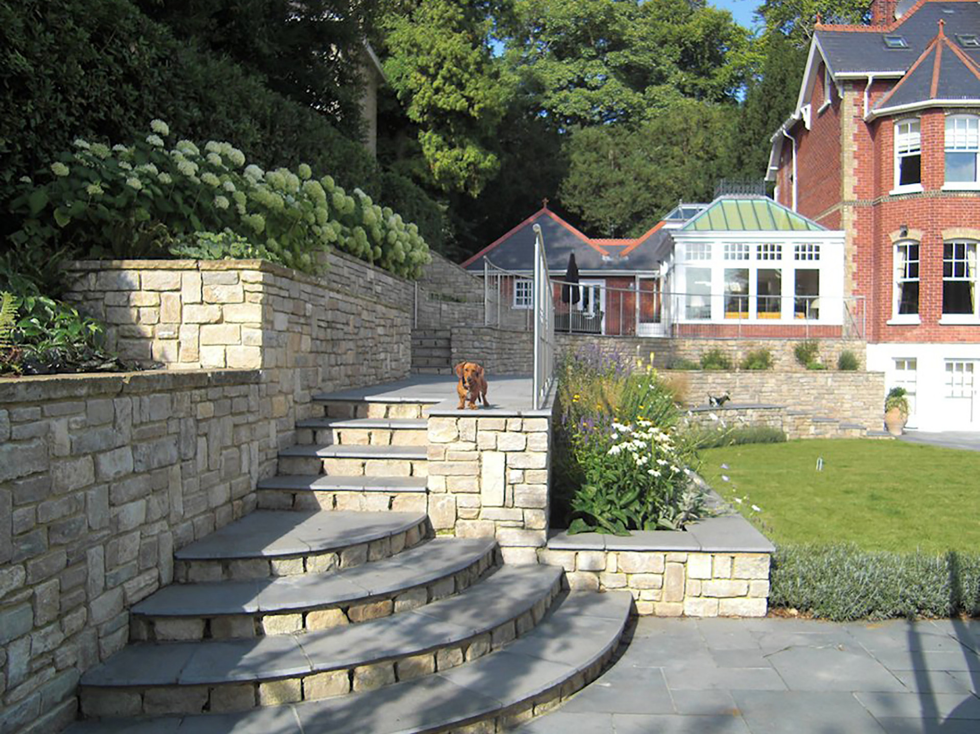 Large sloping garden design in Kenley, Surrey with stone faced retaining walls, semi-circular steps, and stainless steel railings.